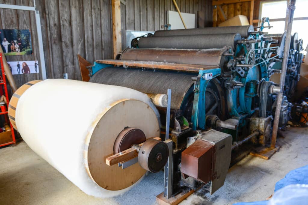 Eine große Walzmaschine in einem rustikalen Raum mit Holzwänden, auf der ein großer Zylinder aus weißem Material gewickelt ist.