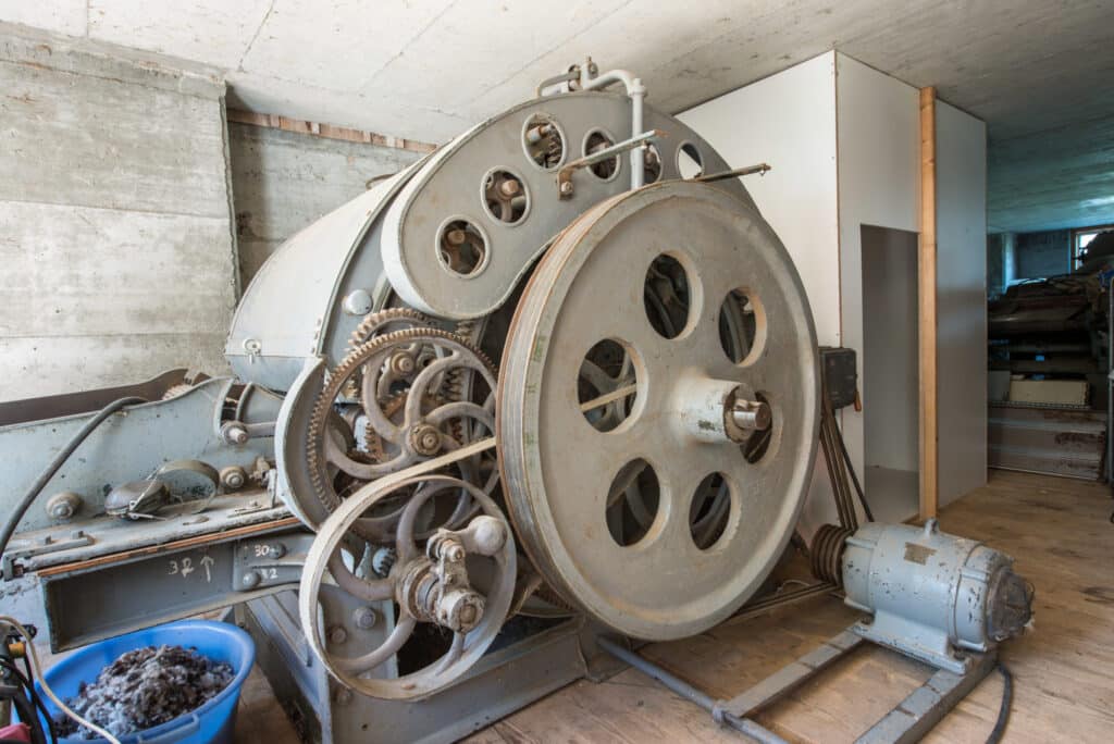 Ein rustikales Gerät mit großen Zahnrädern und Schwungrädern in einem Kellerraum, daneben ein blauer Eimer mit Wolle.
