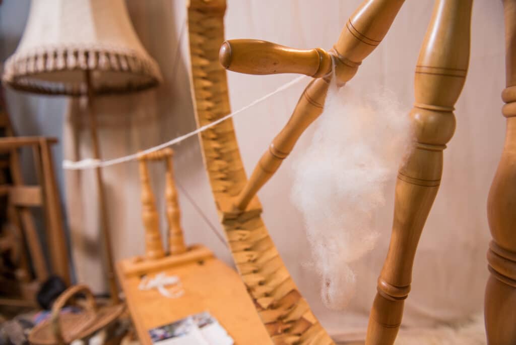 Ein traditionelles Spinnrad aus Holz mit gesponnenem Faden im Fokus, im Hintergrund sind ein geflochtener Korbstuhl und eine Lampe zu sehen.