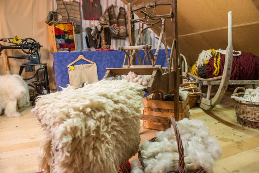 Ein Raum mit handwerklichem Ambiente, in dem Wolle und Filzprodukte verarbeitet werden, mit einem alten Spinnrad, Körben voller Wolle, und farbigen Garnrollen auf einem Regal im Hintergrund.