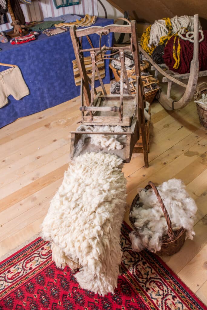 Eine traditionelle Wollbearbeitungsmaschine aus Holz, umgeben von aufgehäufter Wolle und Wollprodukten auf einem hölzernen Boden, mit einem bunten Teppich im Vordergrund.
