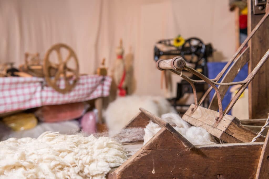 Nahaufnahme einer alten Wollkardiermaschine mit verwirrtem Wollhaufen; im Hintergrund unscharfe traditionelle Spinn- und Webereiausrüstung mit rot-weiß karierten Tischtüchern.