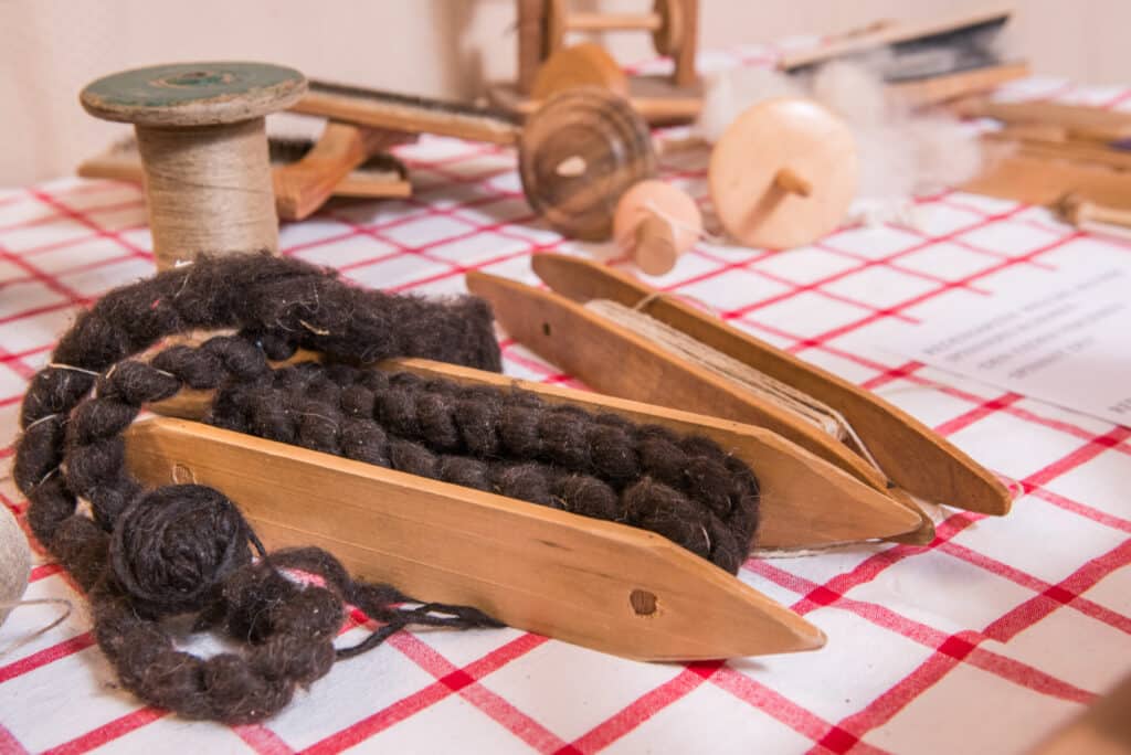 Handwebgerät mit schwarzem Wollgarn auf einem rot-weiß karierten Tischtuch, umgeben von Holzspulen und anderen traditionellen Spinnwerkzeugen.