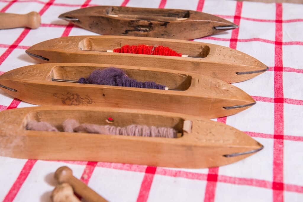 Vier hölzerne Weberschiffchen mit Garn in verschiedenen Farben liegen auf einer rot-weiß karierten Tischdecke.