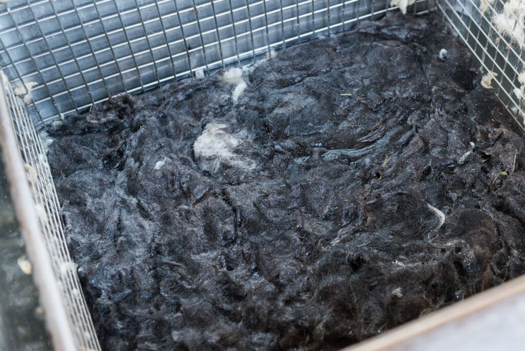 Dunkle, zusammengepresste Wollfasern in einem Drahtkorb mit vereinzelten weißen Stellen.