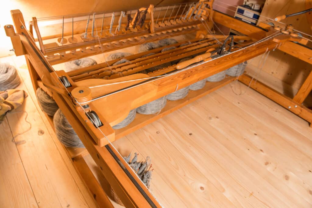 Ein hölzerner Webstuhl mit grauen Garnspulen, aufgestellt auf einem Holzboden in einem Zimmer mit Holzwänden.