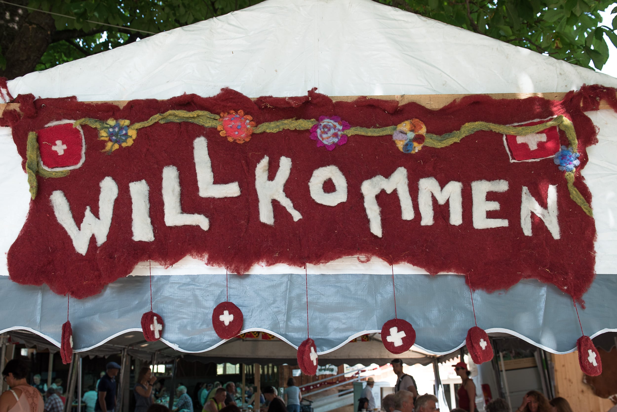Ein handgefertigtes Wollbanner mit der Aufschrift "WILLKOMMEN" in großen, weißen Buchstaben auf einem roten Hintergrund, dekoriert mit Blumenmotiven und Schweizer Kreuzen, ist über einem Zelteingang aufgehängt.