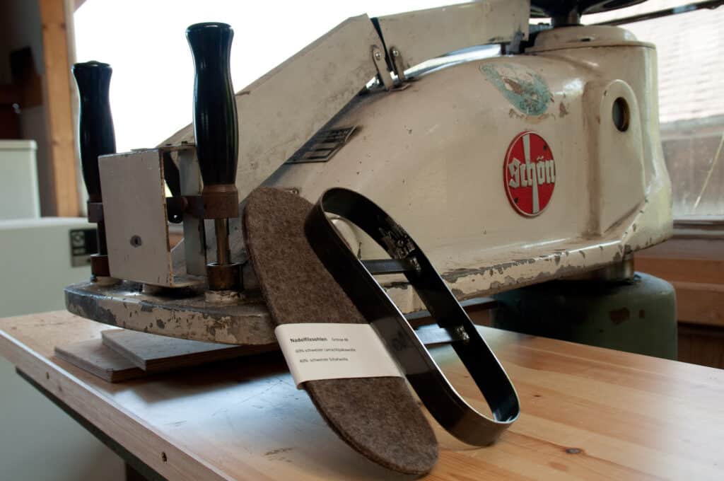 Alte Stanzmaschine mit einem Stück Filzstoff und einem Metall-Schneidrahmen auf einem Holztisch, im Hintergrund ist ein Fenster zu sehen.