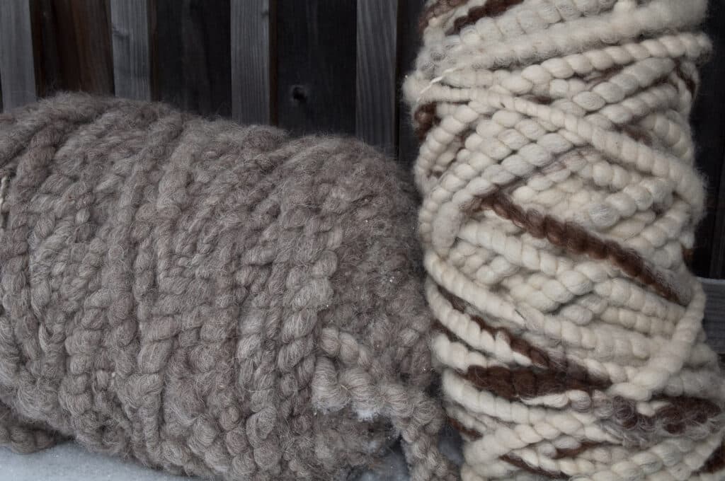 Zwei große Wollknäuel, eins in grauer Farbe und das andere cremefarben mit braunen Streifen, liegen nebeneinander vor einem Holzzaun.