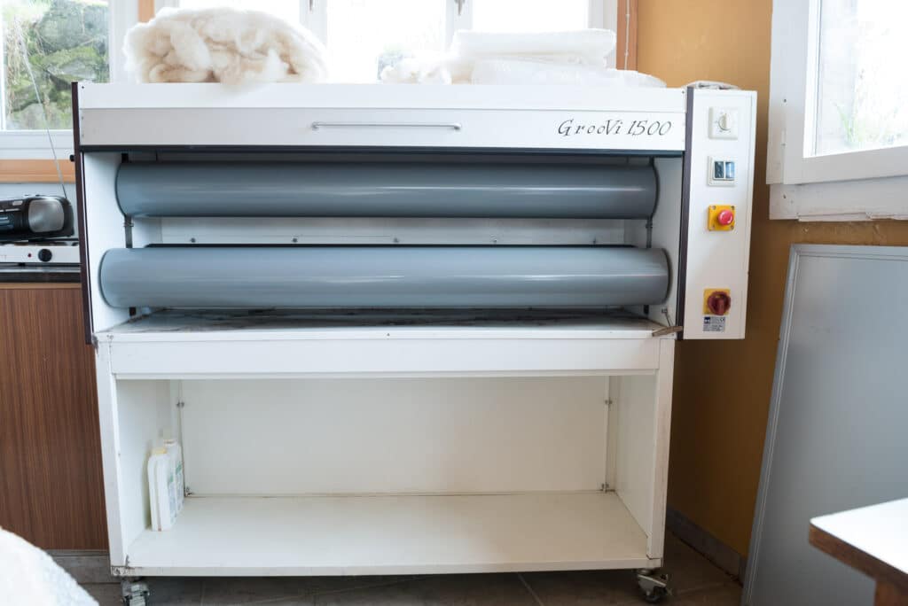 Eine Textilmaschine namens Groovi 1500 mit grauen Walzen, umgeben von Arbeitsflächen, mit weißen Stoffstücken obenauf.