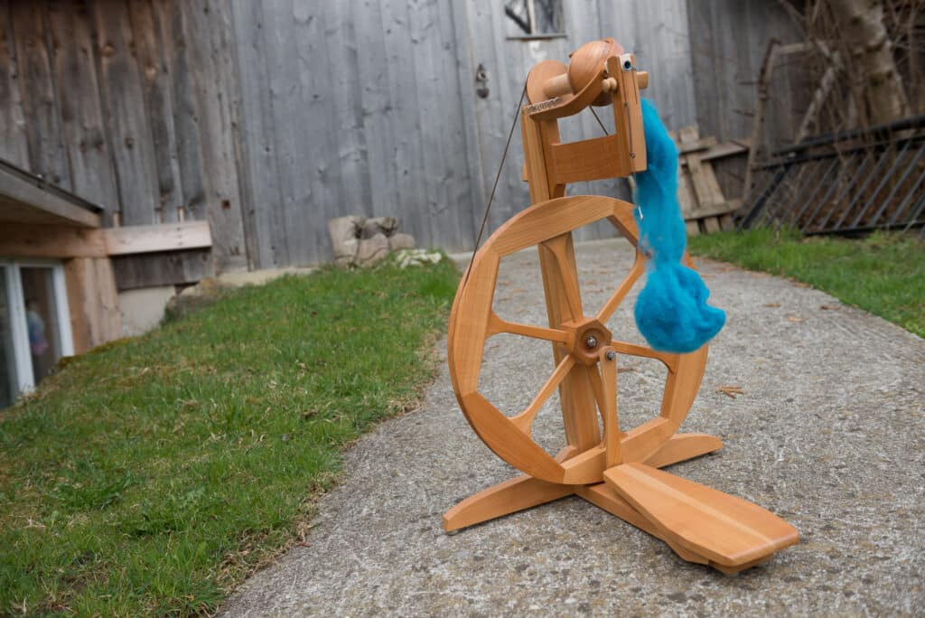 Holzspinnrad mit blauer Wolle auf Steinboden, im Freien vor einer Holzwand platziert.