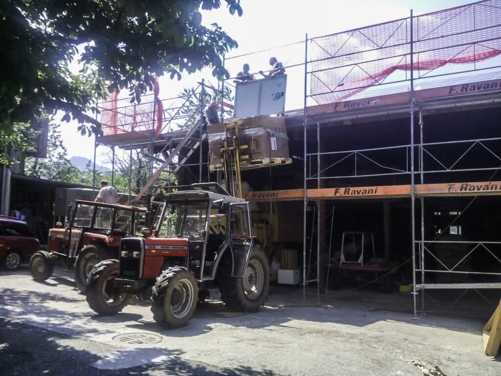 Eine Baustelle mit Gerüst, auf dem mehrere Personen stehen, während ein Traktor einen Gabelstapler nutzt, um Material zu heben. Es gibt zwei weitere Traktoren im Vordergrund.