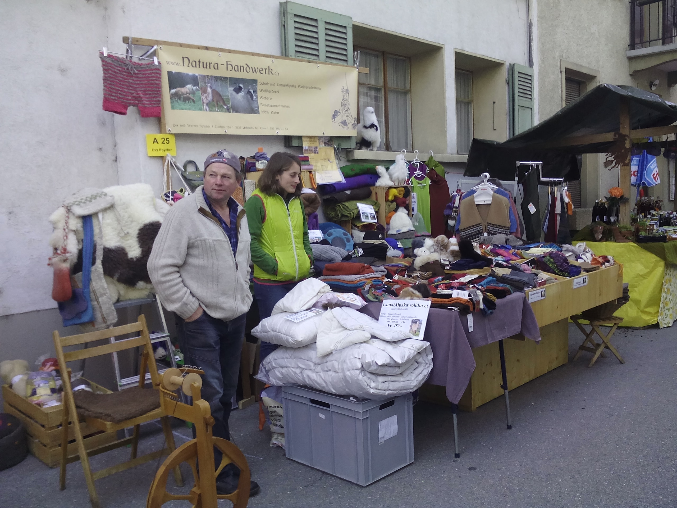 Ein Marktstand mit handgefertigten Textilien. Zwei Personen stehen davor, eine Person trägt eine grüne Jacke. Der Stand bietet Wollprodukte und Alpaka-Waren an, mit sichtbaren Schildern auf den Tischen. Viele farbige Kleidungsstücke sind ausgelegt. Im Hintergrund hängt ein Schild mit der Aufschrift 'Natura-Handwerk'.