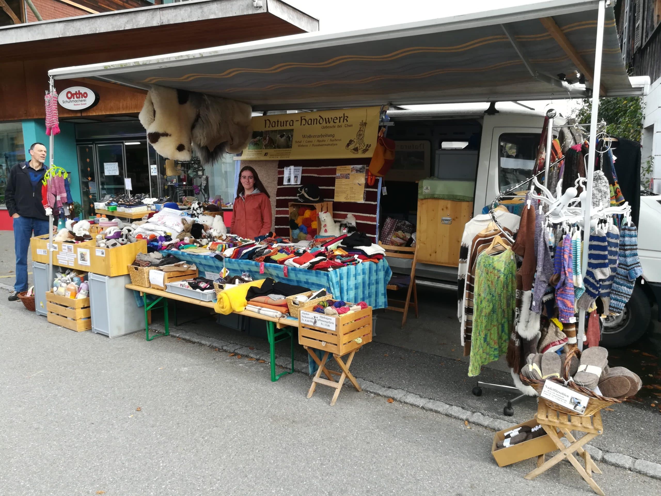 Marktstand mit handgefertigten Produkten aus Wolle, wie Socken und Pullover, die unter einem Vordach präsentiert werden. Ein Mann und eine Frau stehen hinter dem Tisch, auf dem die Waren ausgelegt sind.