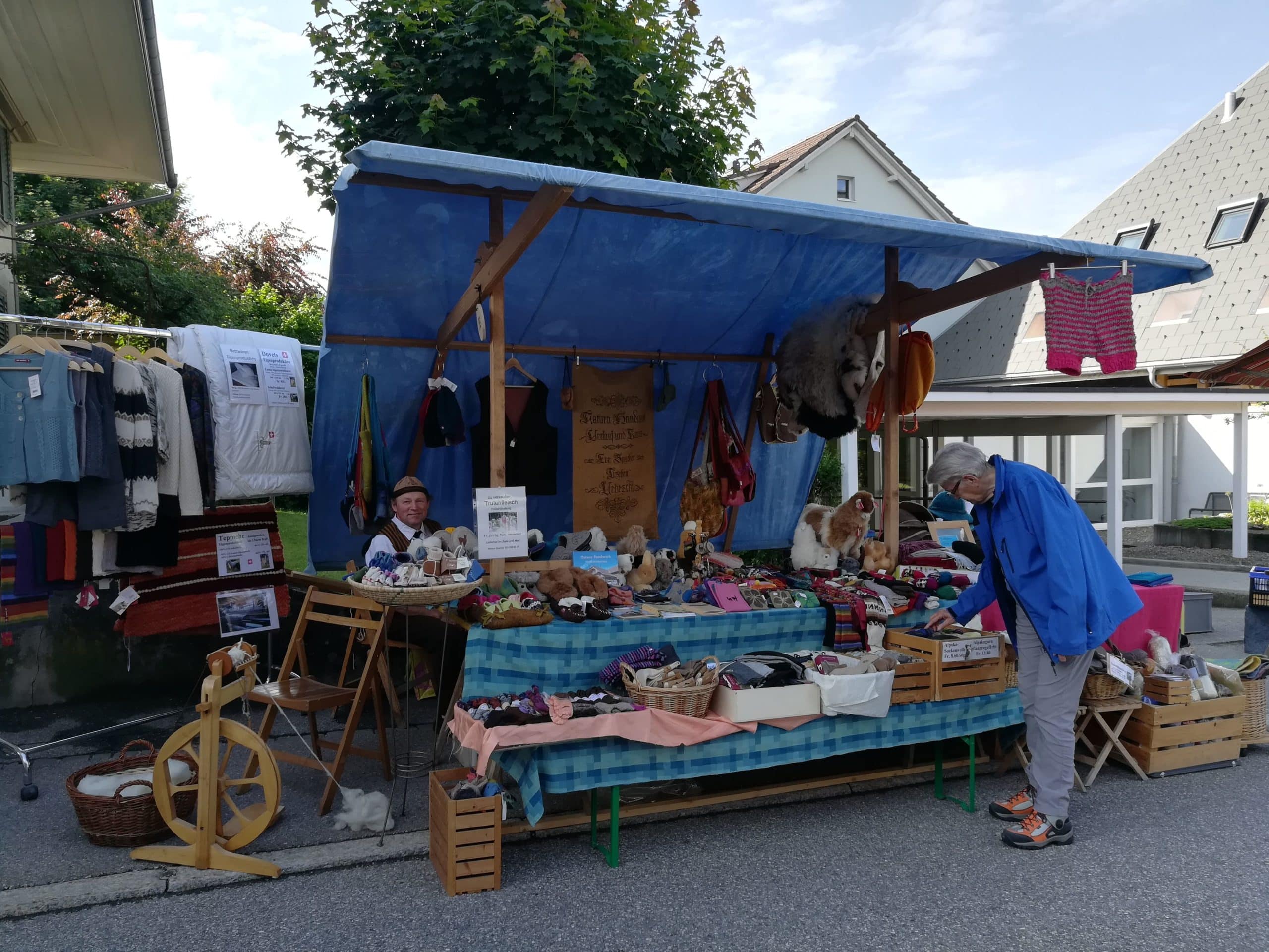 Ein Marktstand mit blauer Markise, an dem ein älterer Mann handgemachte Produkte wie Strickwaren und Stofftiere betrachtet. Ein Verkäufer sitzt hinter dem Tisch, der mit verschiedenen Artikeln bedeckt ist. Der Stand steht auf einer Straße mit Häusern im Hintergrund.
