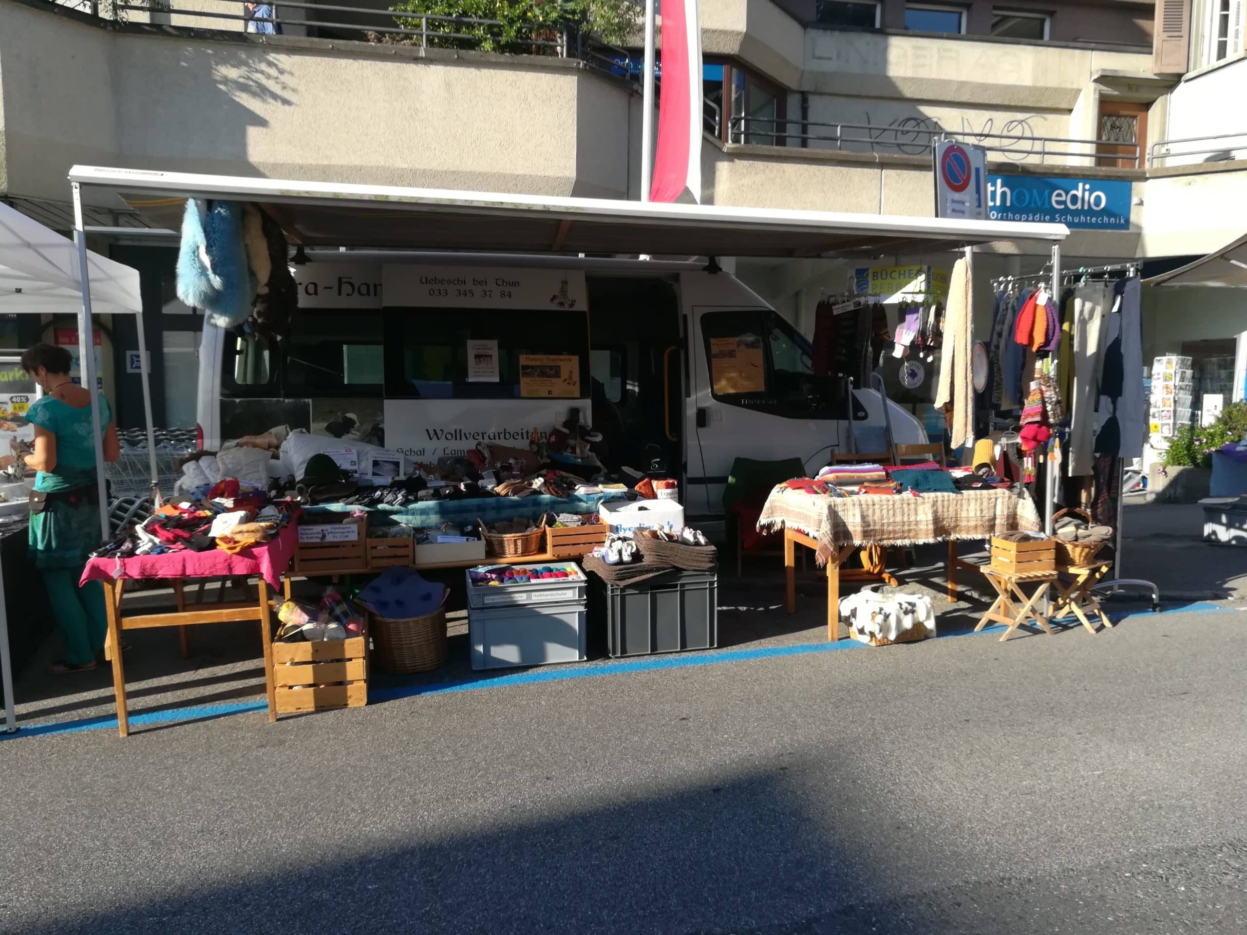 Ein Straßenmarktstand mit einem weißen Lieferwagen im Hintergrund, der handgefertigte Wollprodukte verkauft. Auf Tischen und in Kisten liegen Strickwaren, Schals und andere Textilien ausgestellt. Eine Person steht links neben dem Stand. Im Hintergrund sind Gebäude und ein Schild mit der Aufschrift 'thomedio Orthopädie Schuhtechnik' zu sehen.