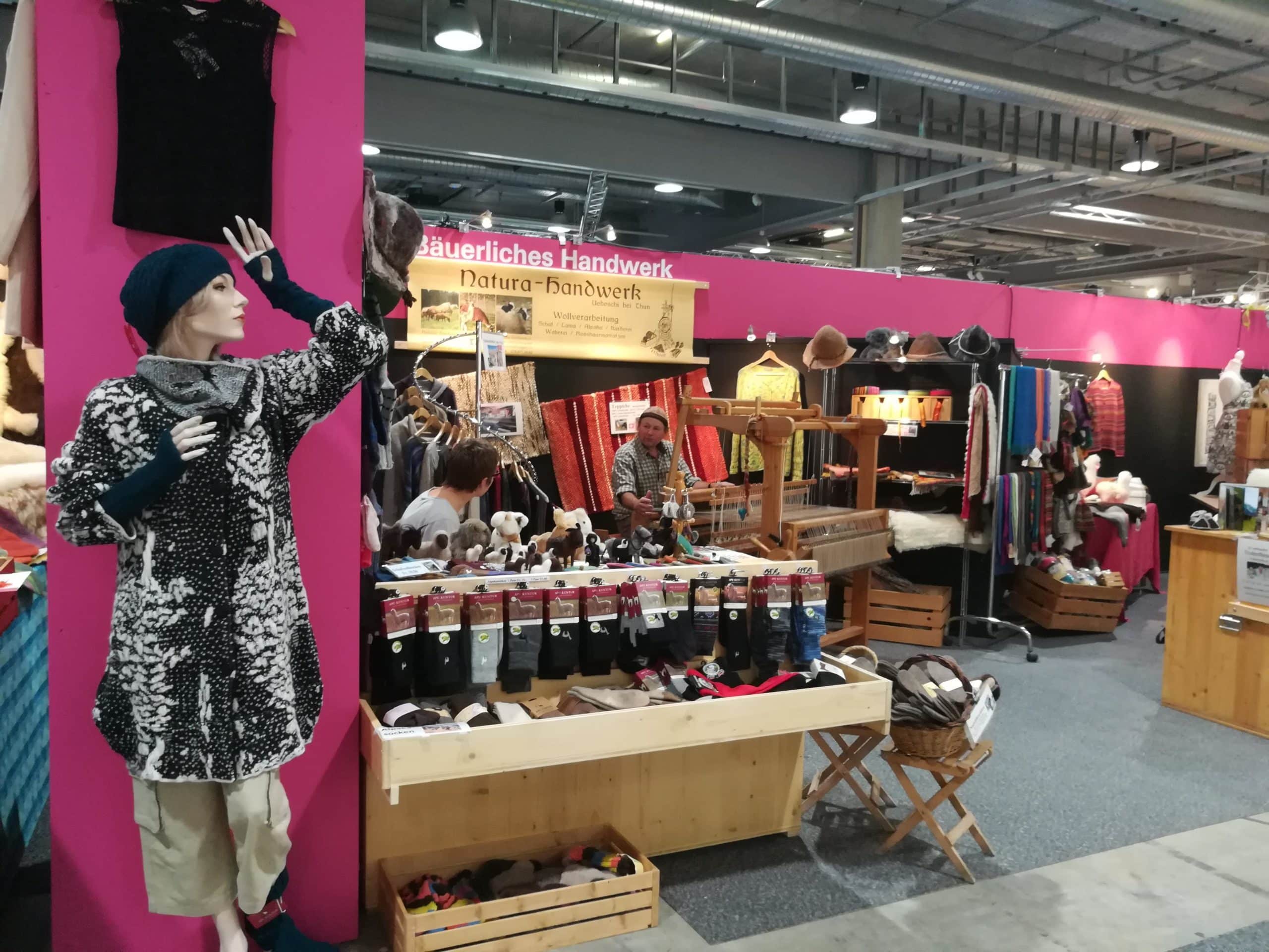 Messestand mit handwerklichen Produkten, darunter Kleidung, Hüte und Accessoires; im Vordergrund eine Schaufensterpuppe mit einem gestrickten Mantel und einem Schal.
