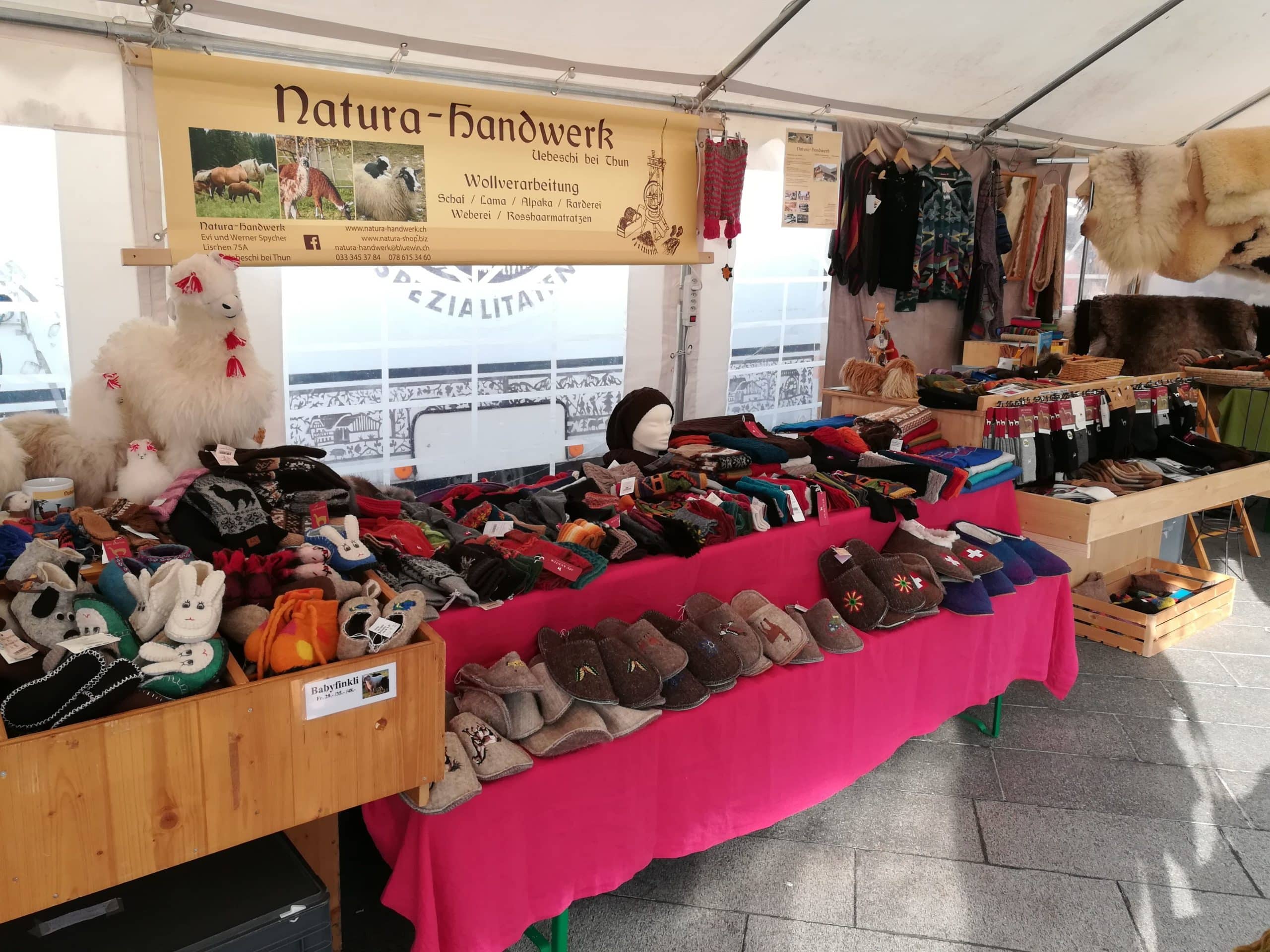 Marktstand mit handgefertigten Wollprodukten wie Filzpantoffeln, Mützen und Spielzeugen, präsentiert auf einem Tisch mit pinker Tischdecke unter einem Zelt mit einem Schild der Marke "Natura-Handwerk".