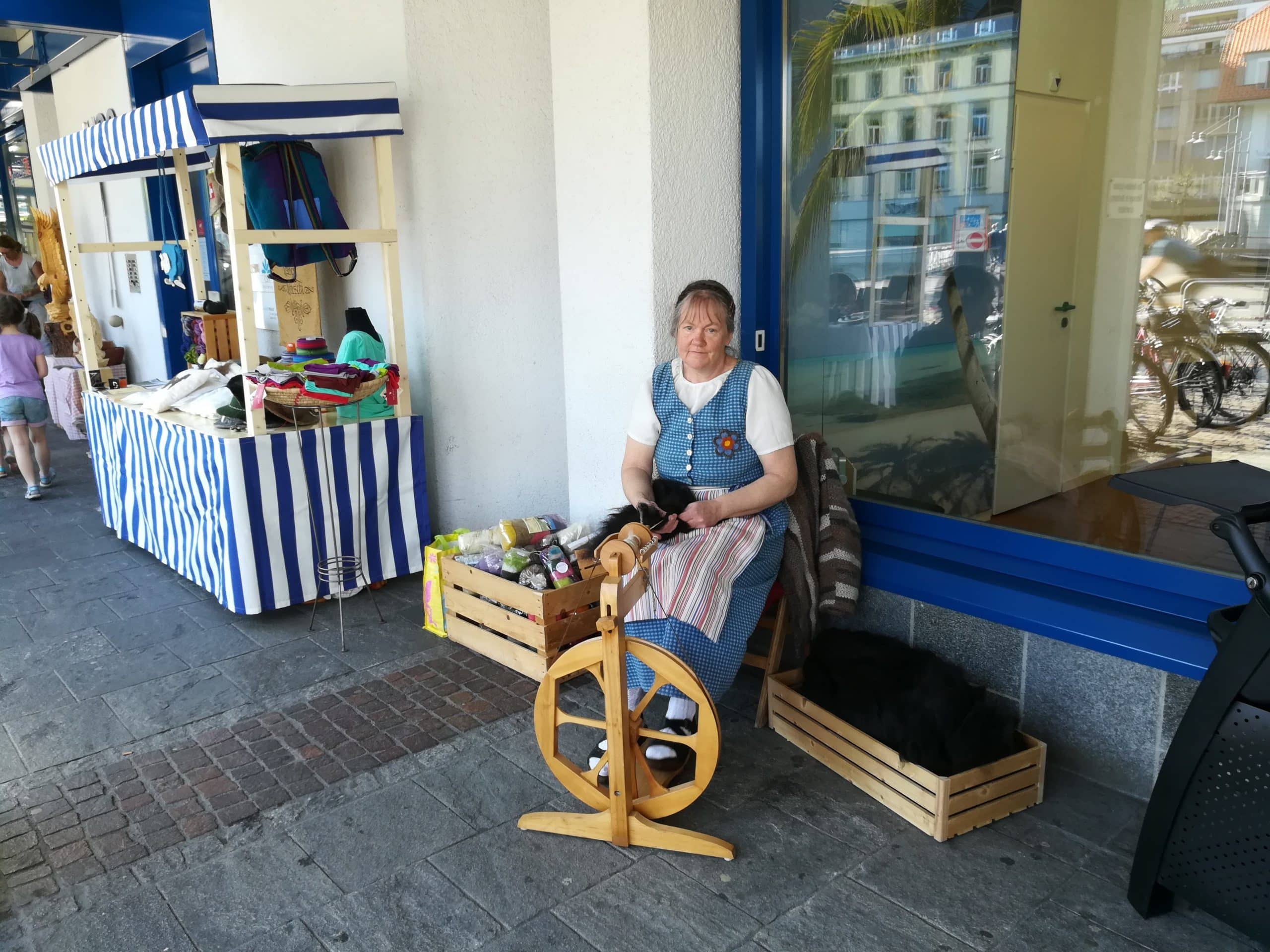 Eine Frau in traditioneller Kleidung sitzt vor einem Laden an einem Spinnrad, daneben ein Marktstand mit blau-weiß gestreiftem Stoff und verschiedenen Waren.