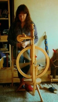 Eine Person sitzt an einem traditionellen Spinnrad in einem Wohnzimmer und spinnt Garn. Im Hintergrund sind Bücherregale und ein Teppich zu sehen.