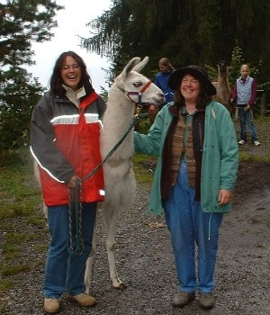Zwei Frauen stehen lächelnd neben einem weißen Lama auf einem Waldweg, beide tragen wetterfeste Kleidung. Im Hintergrund sind Bäume und weitere Personen zu sehen.