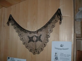 Gehäkeltes Tuch in Dreiecksform mit floralen Mustern, aufgehängt an einer Holzwand, neben Plakaten.