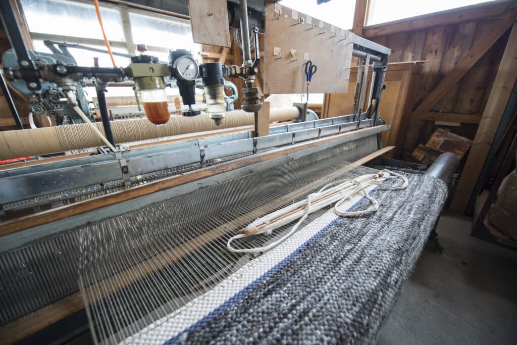 Ein traditioneller Webstuhl in einer Holzhütte, der graue Textilien mit blauen und weißen Akzenten webt, mit verschiedenen Messgeräten und Werkzeugen darüber.