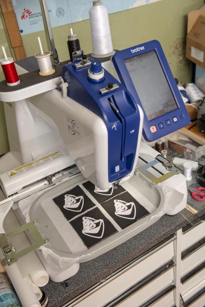 Eine professionelle Stickmaschine mit einem blauen Display fertigt vier weiße Stickmotive auf schwarzen Stoffstücken an. Verschiedene Fadenspulen in Rot, Weiß und Schwarz sind oben auf der Maschine angebracht, und die Umgebung ist eine Werkstatt mit weiteren Materialien und Werkzeugen im Hintergrund.