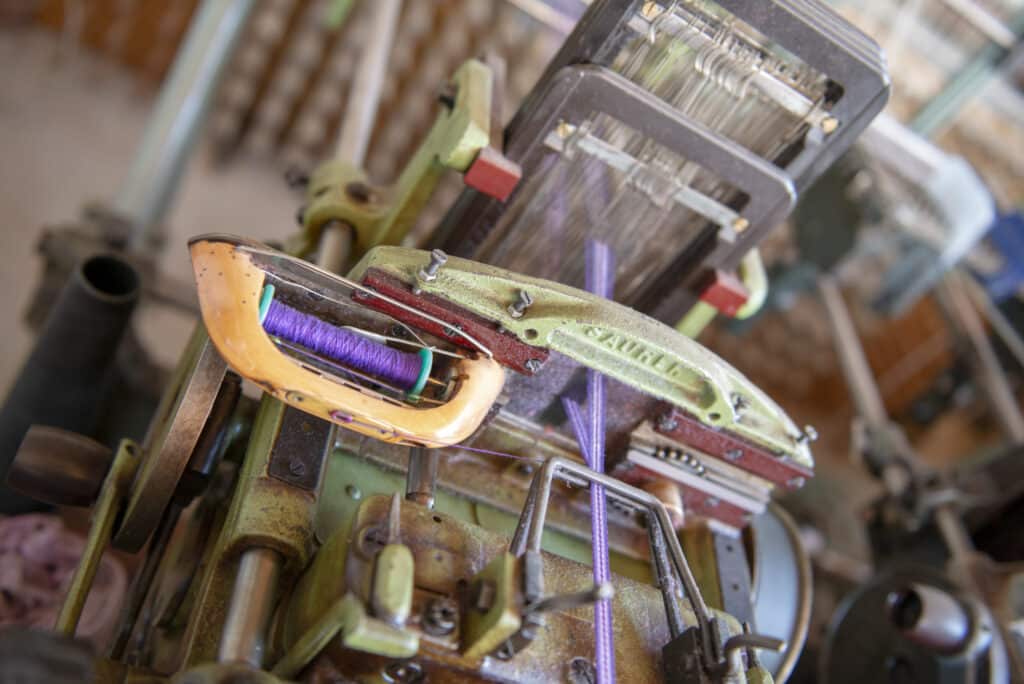 Nahaufnahme einer Webmaschine mit einer Spule aus violettem Garn in einem gelben Fachwerk, umgeben von metallischen Hebeln und Zahnrädern.