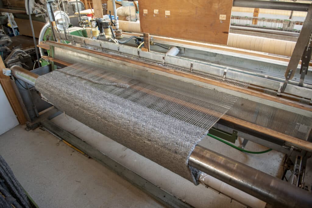 Webstuhl in einer Textilfabrik mit gewebtem Stoff, der aus der Maschine herauskommt, umgeben von mechanischen Teilen und Steuerinstrumenten.