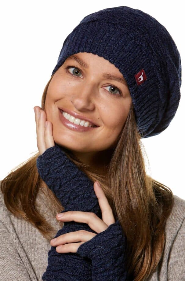 Eine lächelnde Person trägt eine dunkelblaue Strickmütze und passende fingerlose Handschuhe, zur Seite geneigt, mit einer Hand am Kinn.