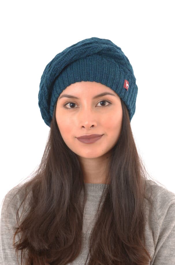 Frau mit langen dunklen Haaren trägt eine blaue Strickmütze und einen grauen Pullover vor einem weißen Hintergrund.