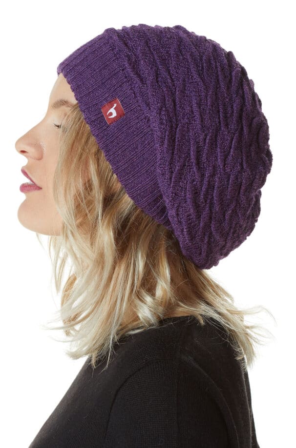 Eine Frau mit welligem blondem Haar trägt eine lila Strickmütze mit einem kleinen, roten Etikett an der Seite, im Profil fotografiert vor einem weißen Hintergrund.