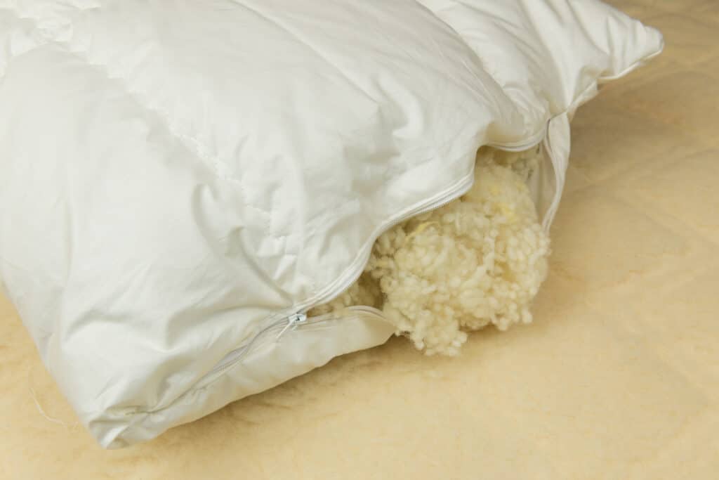 Ein weißes Kopfkissen mit geöffnetem Reißverschluss, aus dem Schafwolle herausragt, liegt auf einem beige-farbenen Hintergrund.