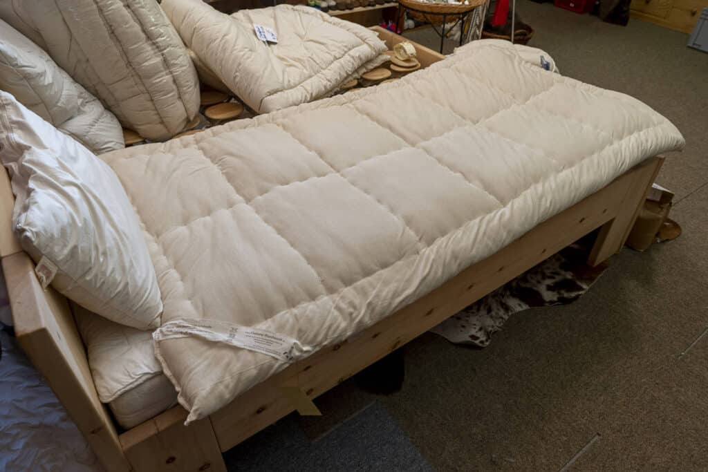 Eine Matratze mit einer weißen gepolsterten Auflage auf einem Holzrahmenbett, umgeben von mehreren zusammengeklappten Bettdecken.