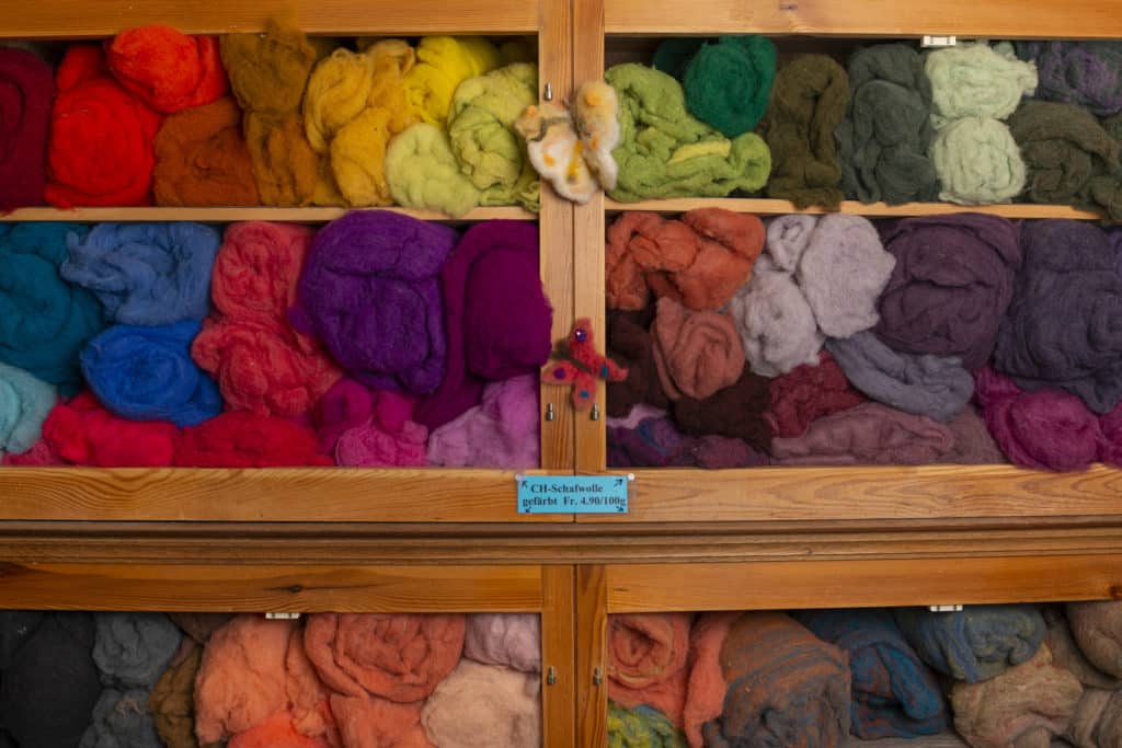 Ein Holzregal mit verschiedenfarbiger Schafwolle, ordentlich nach Farben geordnet, darunter ein Preisschild mit 4.90 Franken pro 100 Gramm.