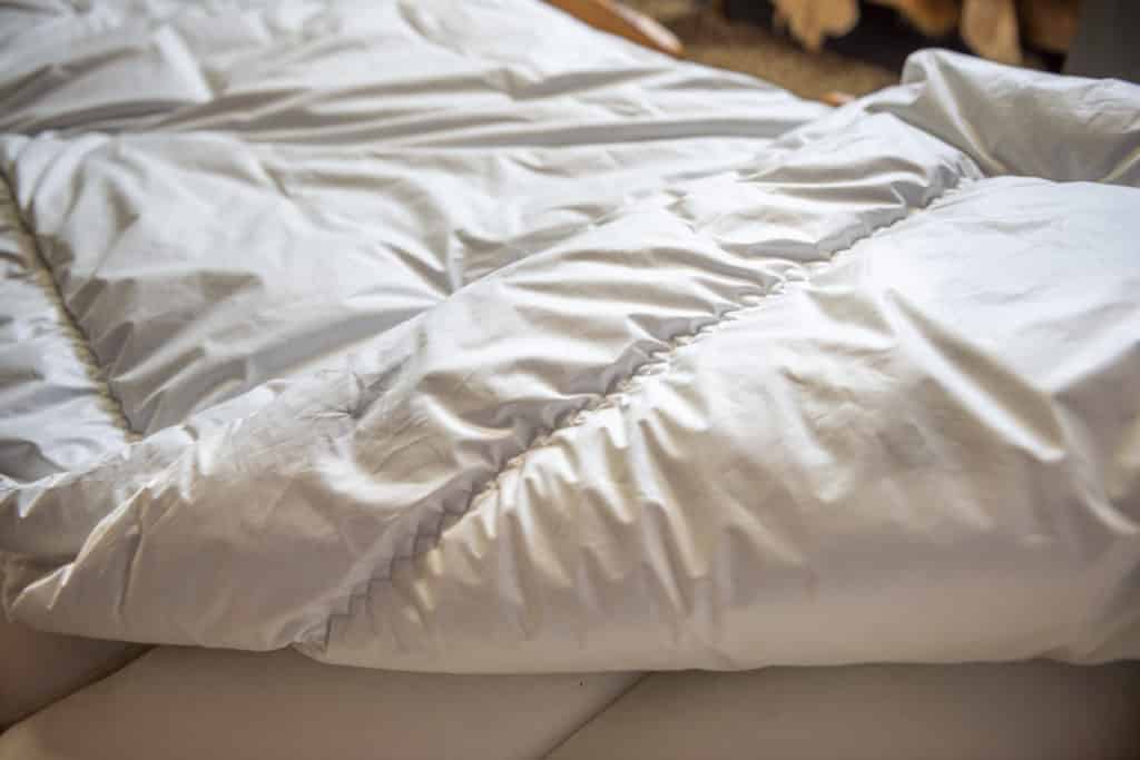 Eine weiße Daunendecke auf einem Bett, leicht zerknittert und in einem wohnlichen Ambiente fotografiert.