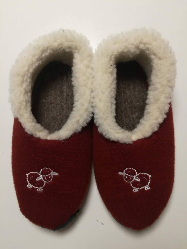 Ein Paar rote Hausschuhe mit weißem Futter und einem gestickten Schafmotiv auf jedem Schuh, auf einem hellen Hintergrund.