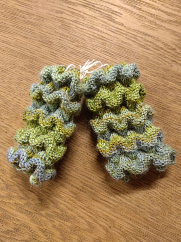 Gestrickte, wellenförmige Handstulpen in Grün- und Blautönen auf einem Holztisch.
