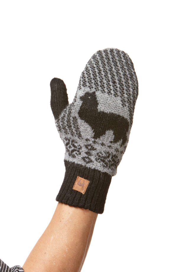 Eine gestrickte Fausthandschuh mit einem schwarzen Alpakamuster auf grauem Hintergrund, getragen an einem Arm, mit einem braunen Etikett am Bündchen.