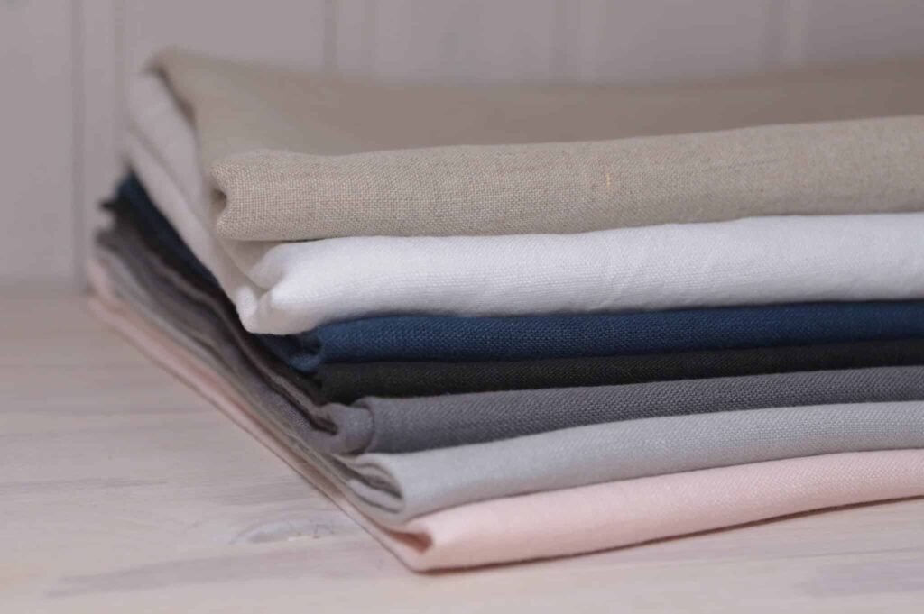 Ein Stapel gefalteter Stoffe in verschiedenen Farben, darunter Beige, Weiß, Dunkelblau, Schwarz, Grau und Rosa, liegt auf einem hellen Holztisch.