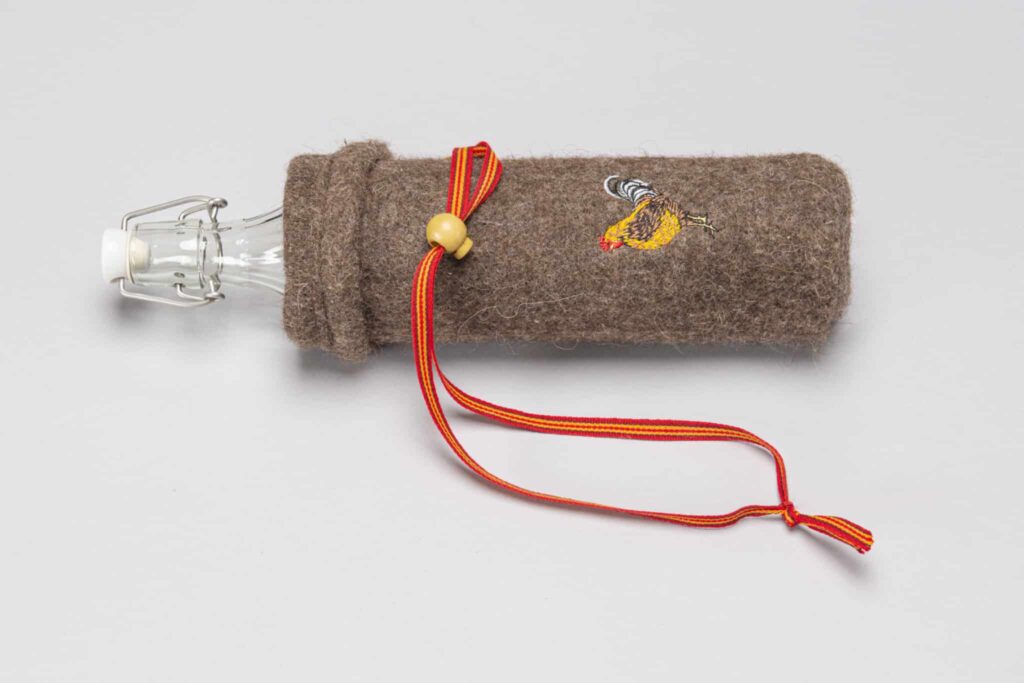 Eine Glasflasche mit Bügelverschluss ist in eine braune Filzhülle eingewickelt, die mit einem gestickten Hahnmotiv und einer rot-gelben Kordel verziert ist.