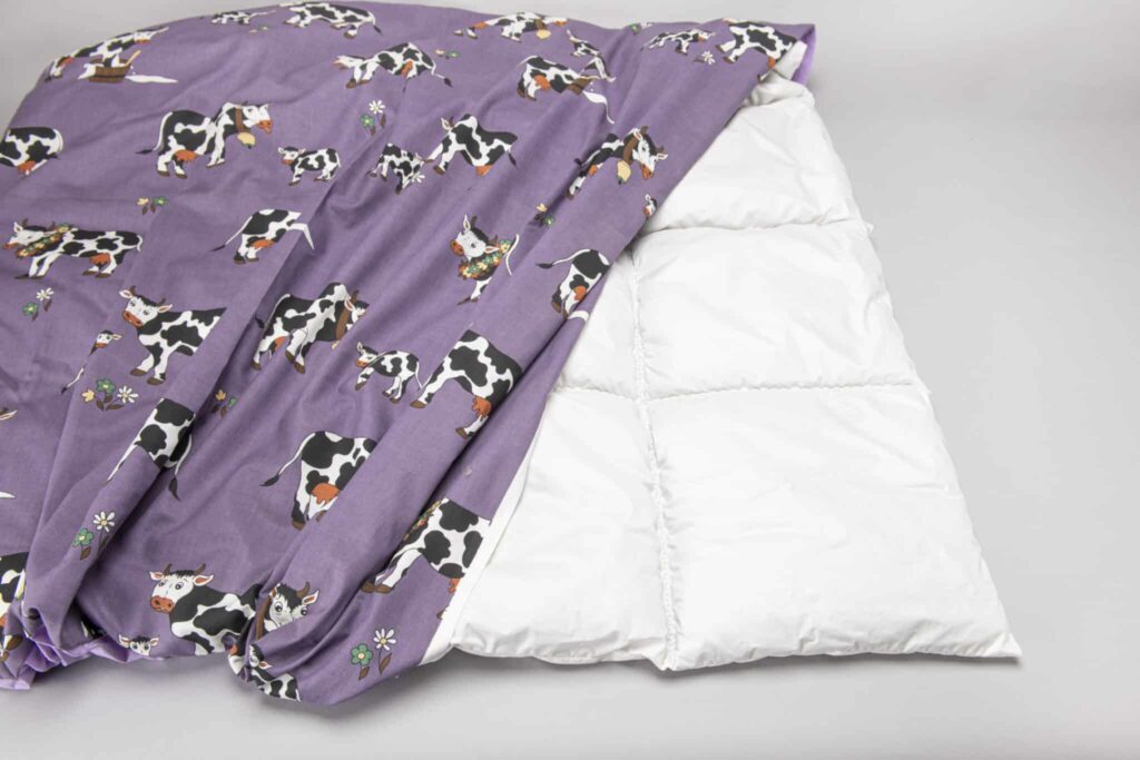 Eine Bettdecke mit einem lila Bezug, der mit Kuhmotiven und kleinen Blumen bedruckt ist, liegt halb über einer weißen Steppdecke.