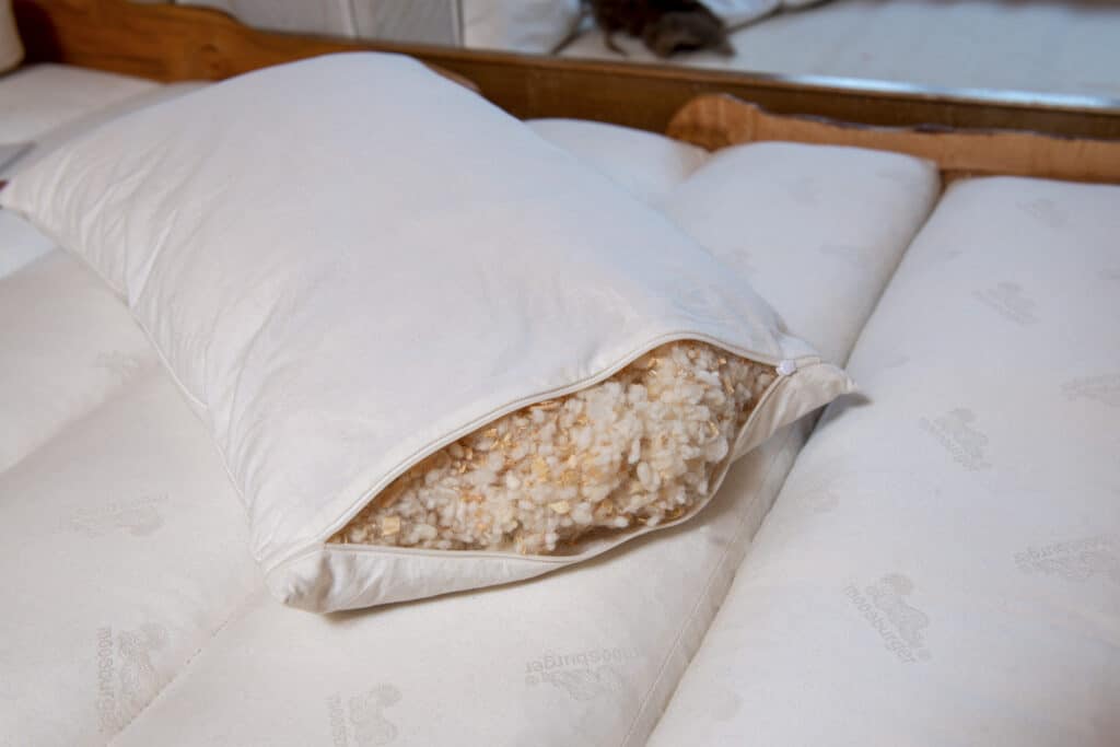 Ein Kissen aus weißem Stoff liegt auf einer Matratze und ein Teil des Innenfutters aus weichem, wollähnlichem Material ist durch einen offenen Reißverschluss sichtbar.