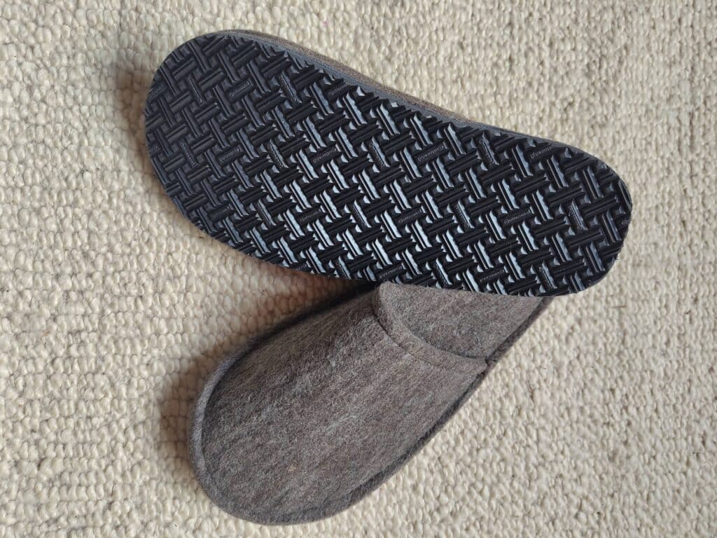 Graue Filzpantoffeln mit strukturierter schwarzer Sohle auf einem beigen Teppich.