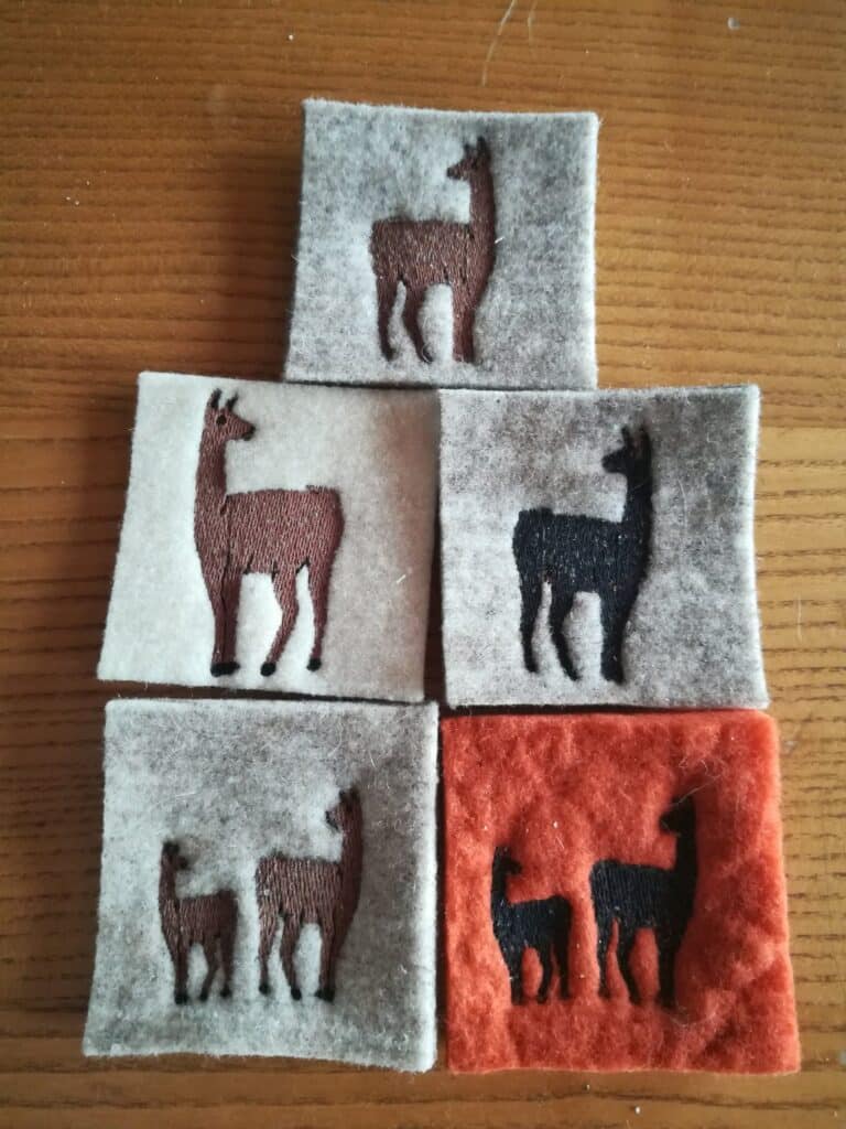 Fünf quadratische Filzstücke in verschiedenen Farben mit aufgestickten Lama-Motiven, die ein pyramidales Muster bilden, auf einer Holzoberfläche arrangiert.