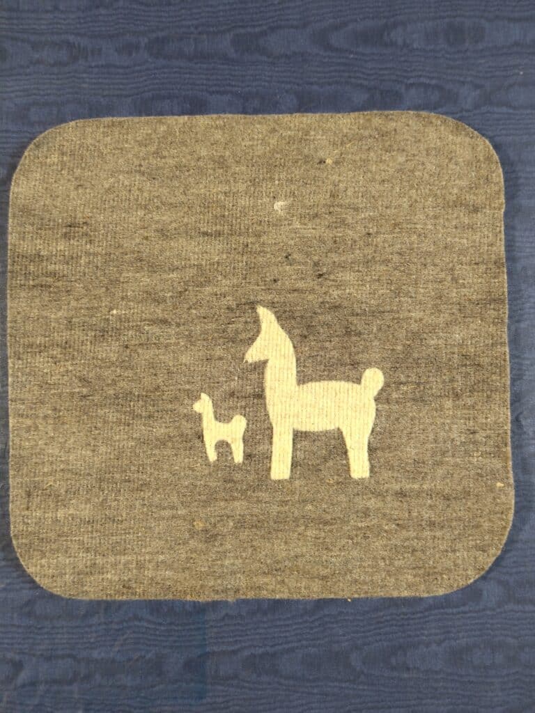 Zwei stilisierte Llama-Silhouetten, eine große und eine kleinere, auf einem grauen rechteckigen Stoff vor blauem Hintergrund.