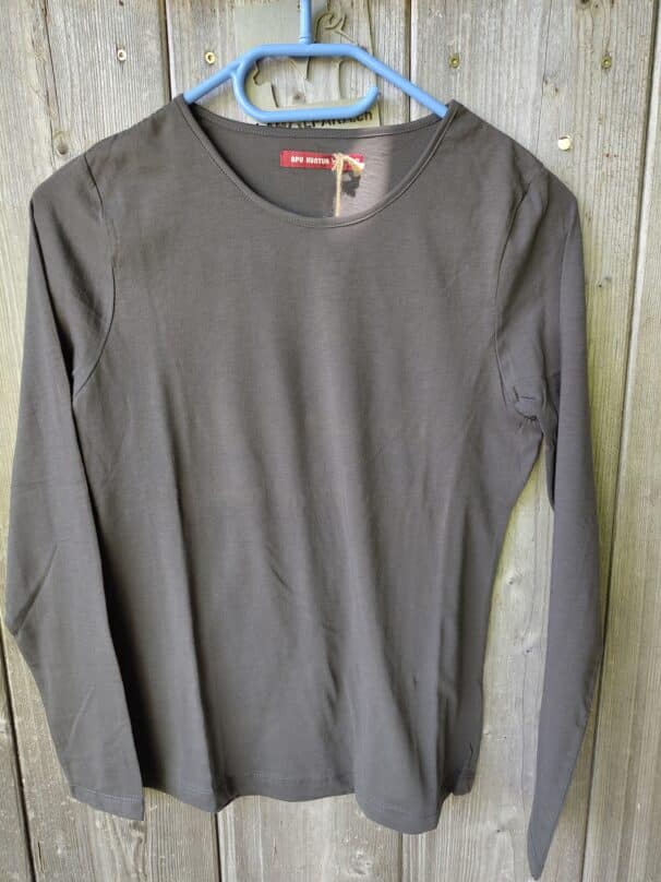 Graues Langarmshirt mit Rundhalsausschnitt auf einem blauen Kleiderbügel vor einem Holzzaun.