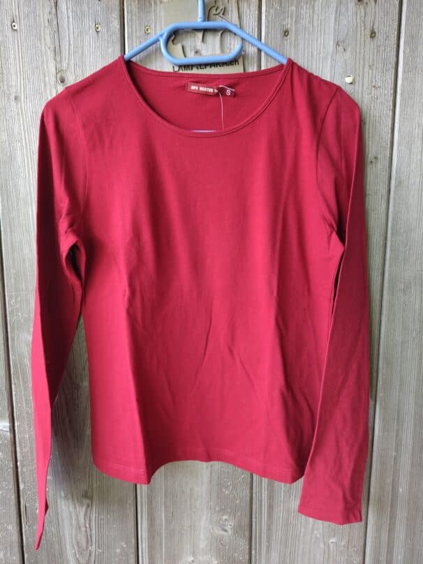 Ein rotes Langarmshirt in Größe S hängt an einem blauen Kleiderbügel vor einem Holzhintergrund.