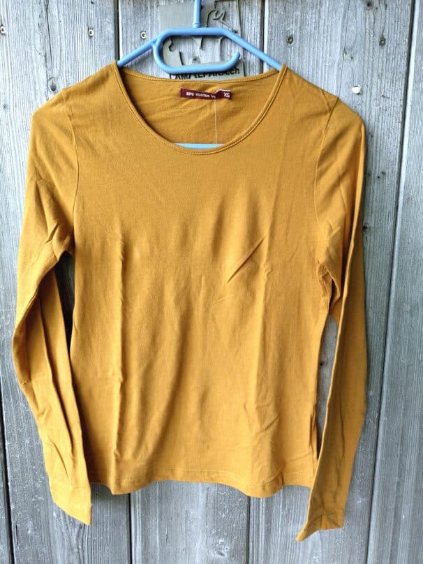 Ein ockerfarbenes Langarmshirt aus Baumwolle, Größe XS, hängt an einem blauen Bügel vor einer grauen Holzwand.